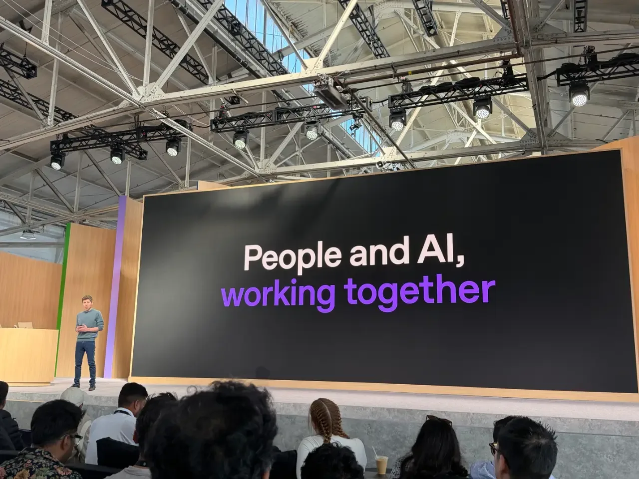 Sam Altman stands on a conference stage before a seated audience, beside a large screen reading “People and AI, working together.”